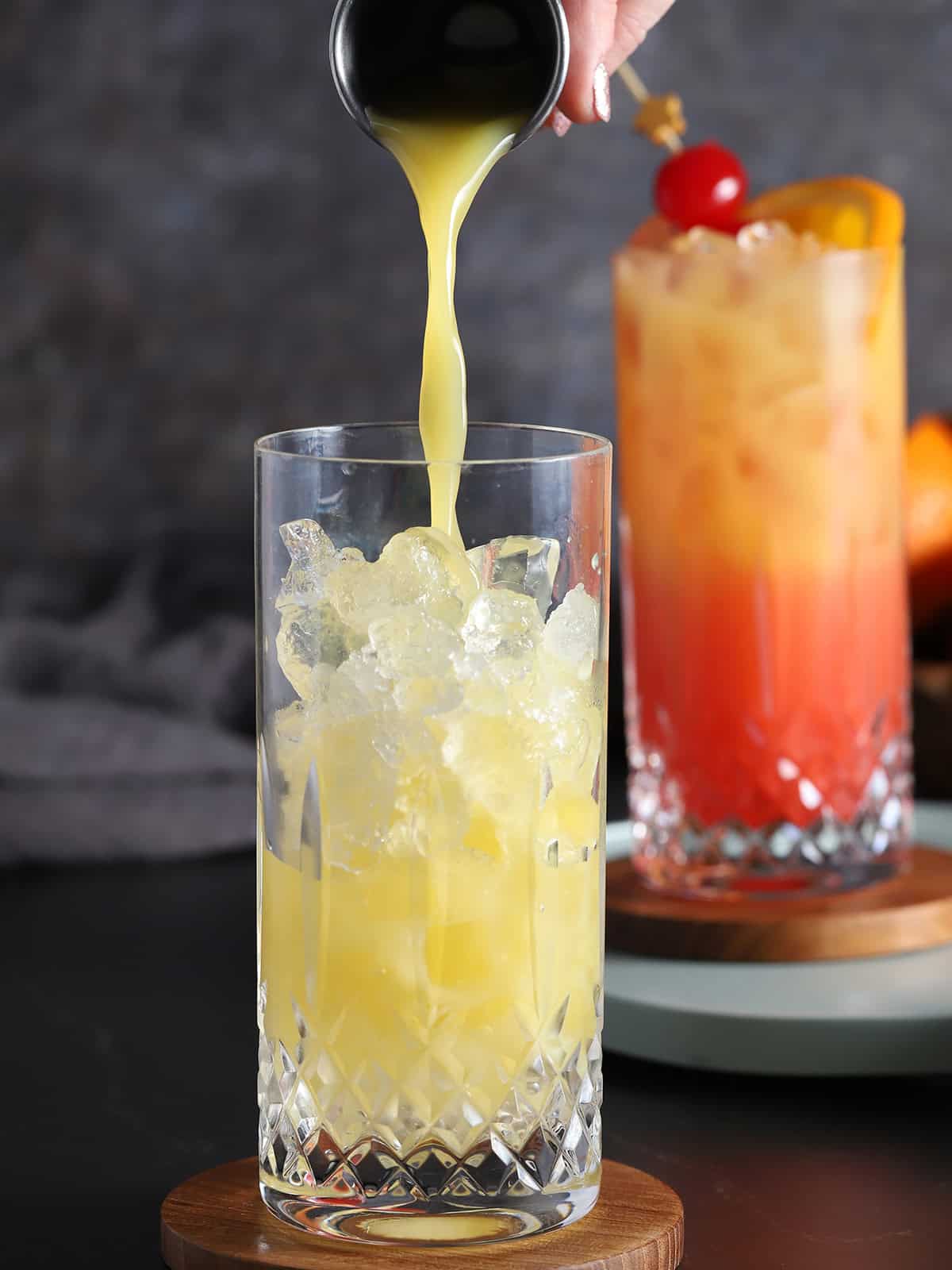 A hand pouring orange juice from a jigger into a tall glass filled with ice.