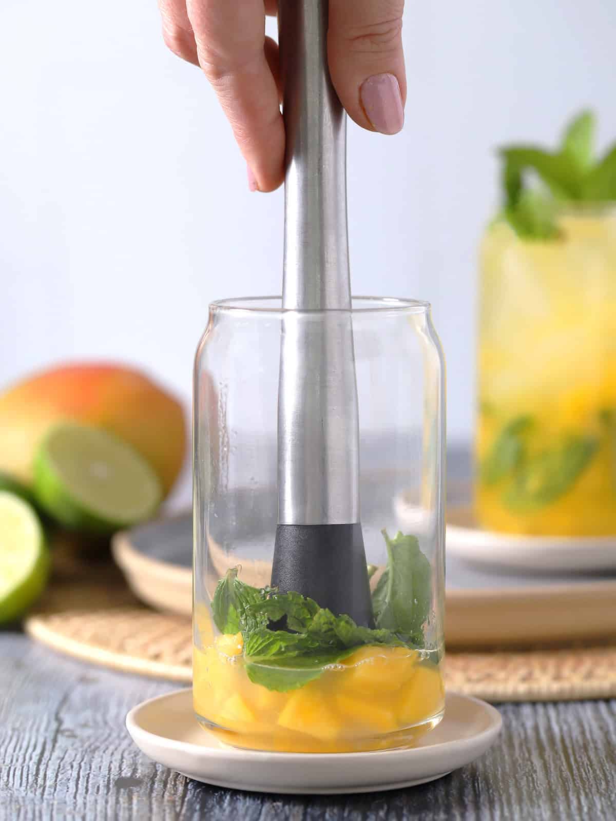 A hand placing a muddler into a glass of mango, lime, and mint.