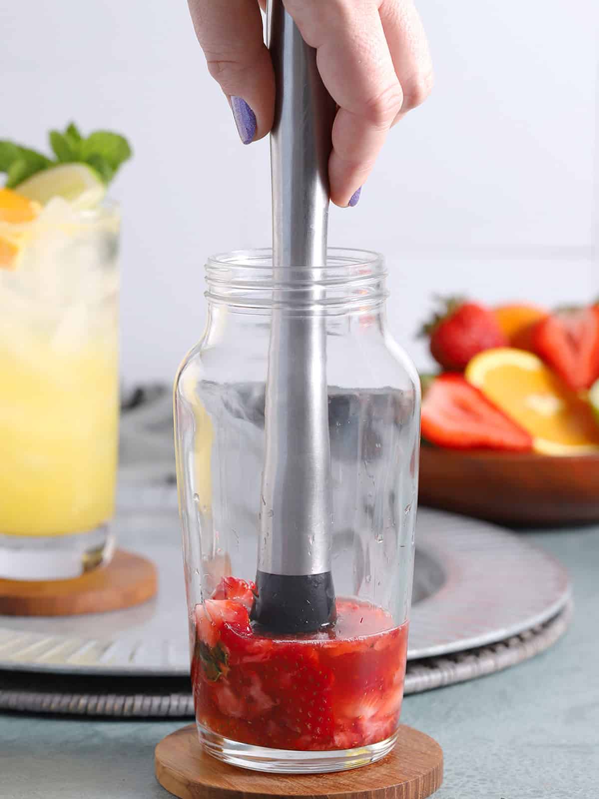A hand muddling berries, mint and syrup in a cocktail shaker.