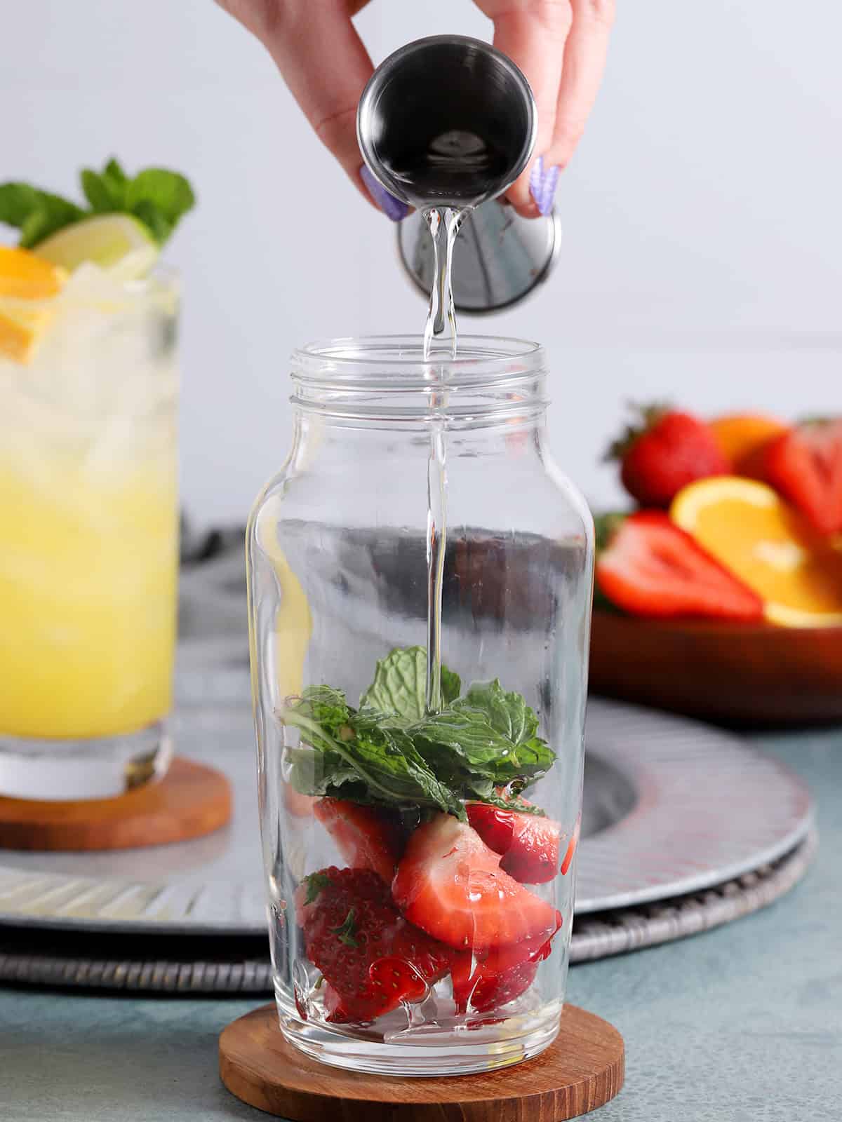 A hand pouring simple syrup from a cocktail jigger into a glass with mint and berries.