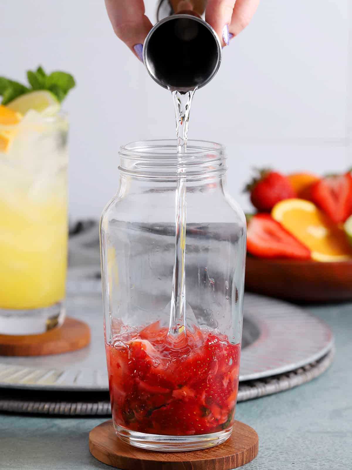 Tequila being poured into a cocktail shaker with berries, mint, and syrup.