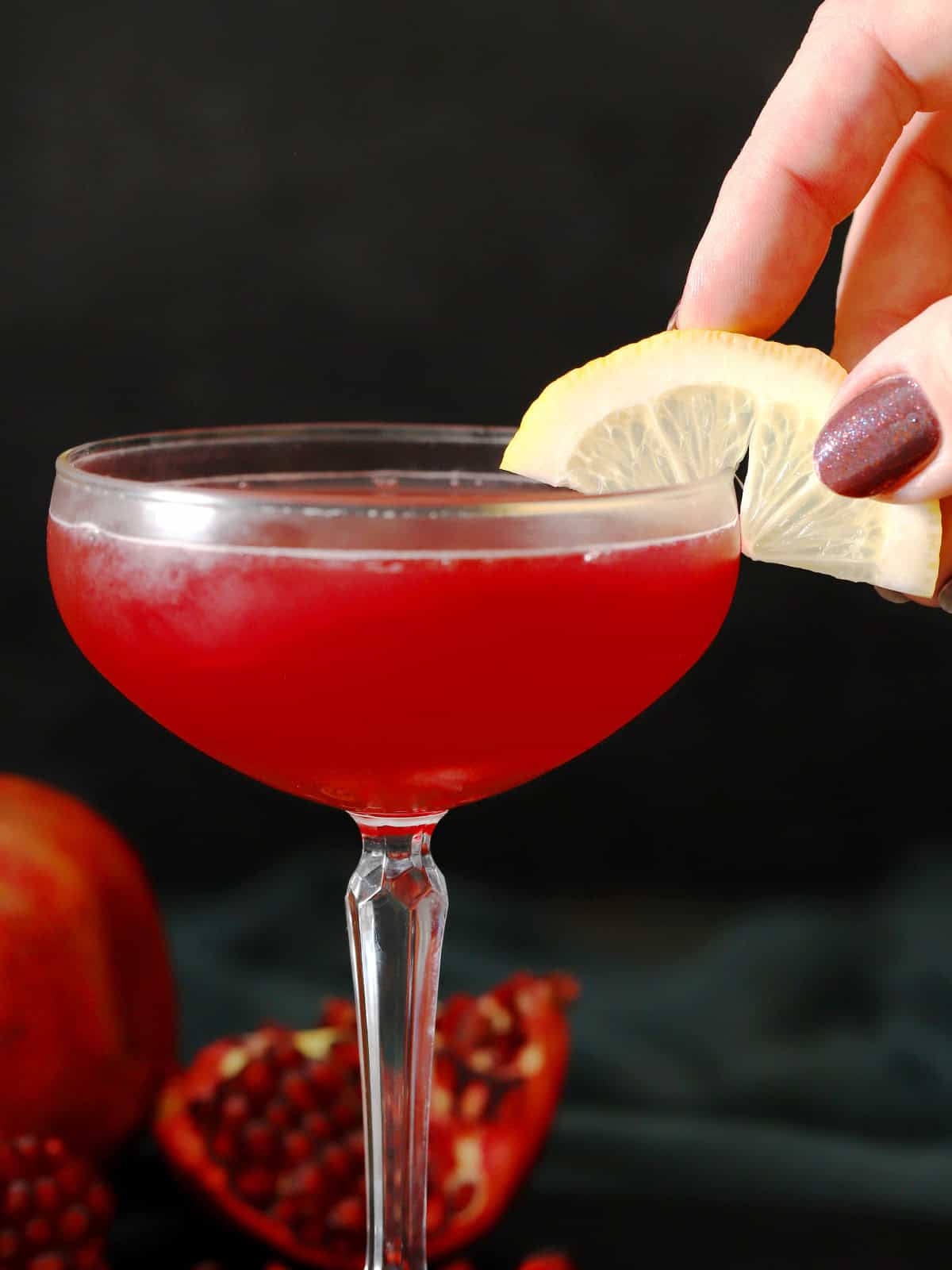 A hand placing a slice of lemon onto the side of a coupe glass filled with pomegranate martini.