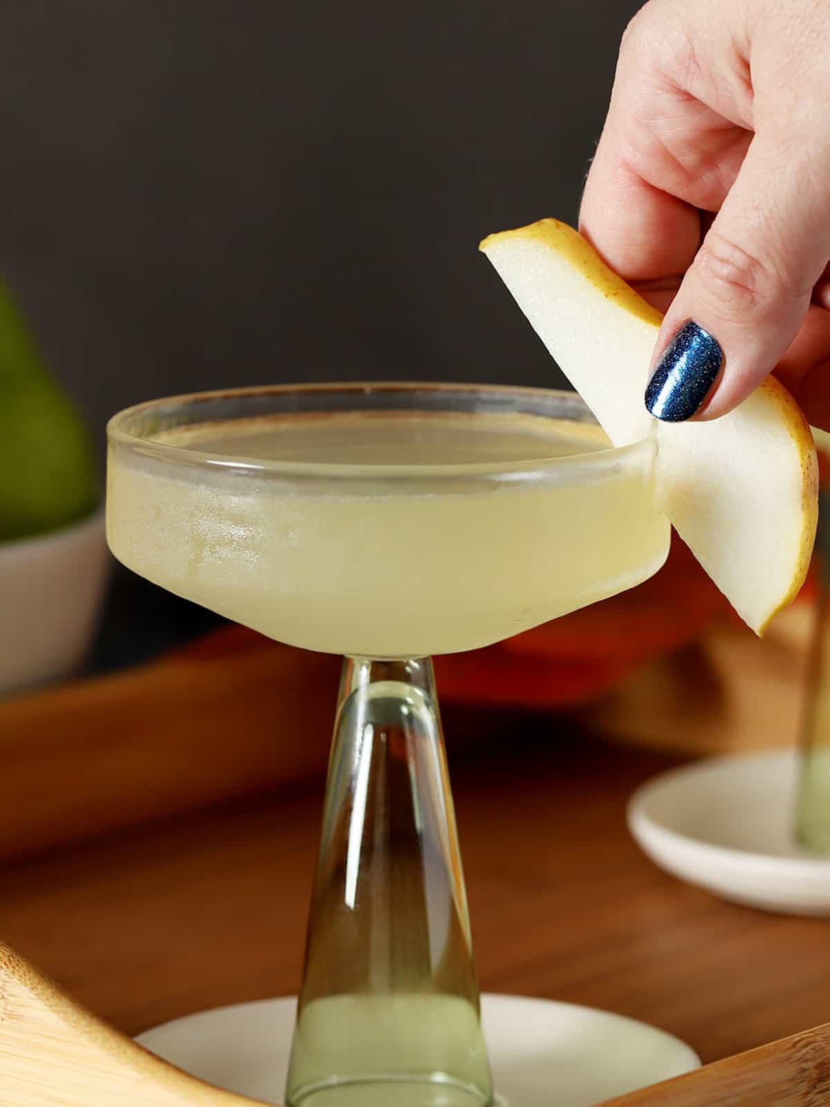 A hand placing a slice of fresh pear onto the side of a coupe glass filled with pear martini.