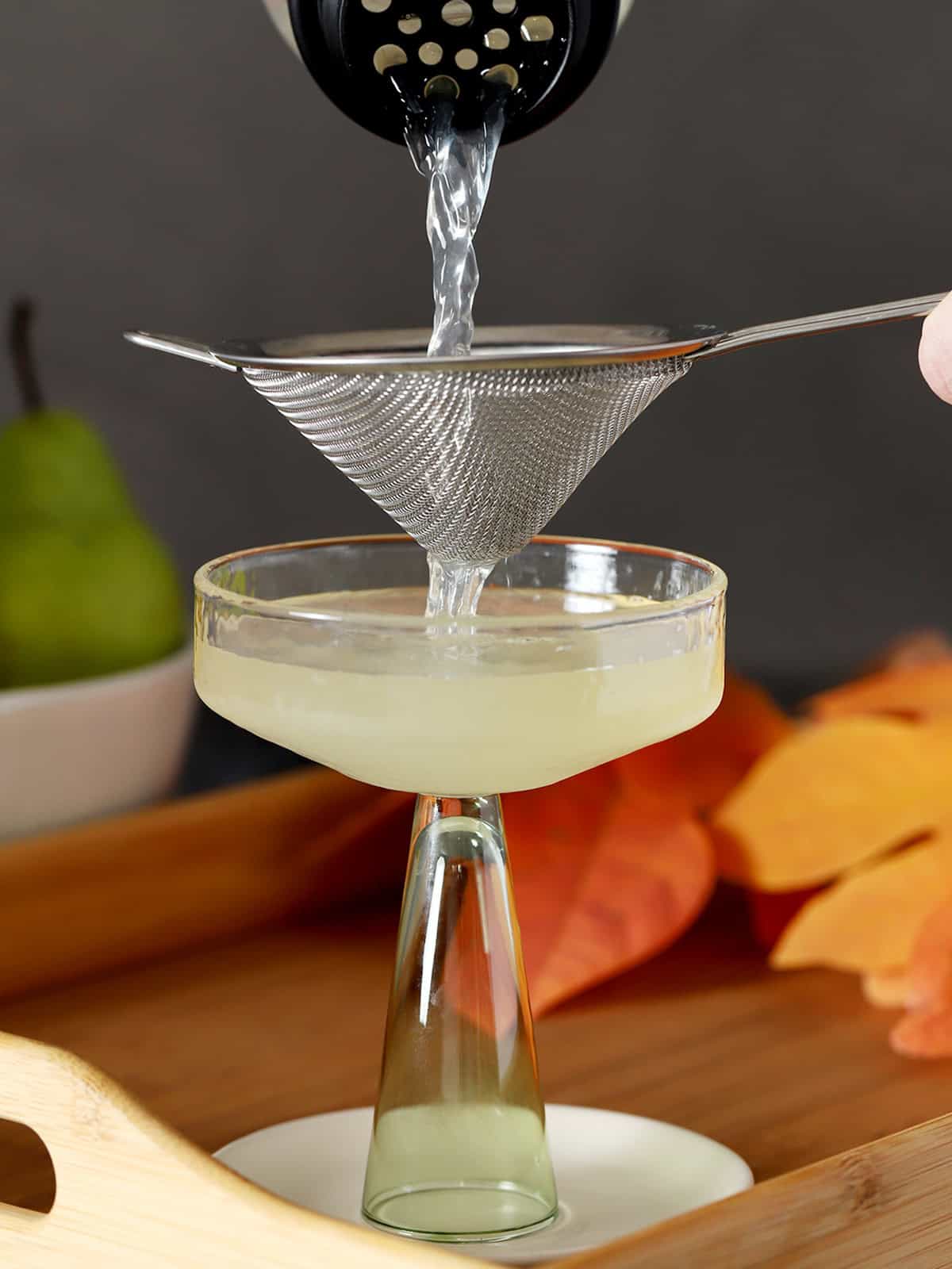 Pear martini being poured from a cocktail shaker, through a fine mesh sieve into a coupe glass.
