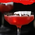 A coupe glass of red Vampire's Kiss cocktail with a black sugar rim, garnished with white plastic vampire teeth.