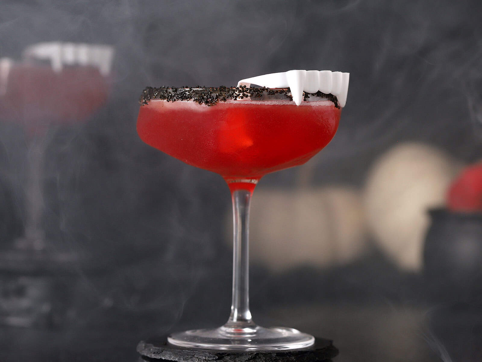 A coupe glass of red Vampire's Kiss cocktail with a black sugar rim, garnished with white plastic vampire teeth.