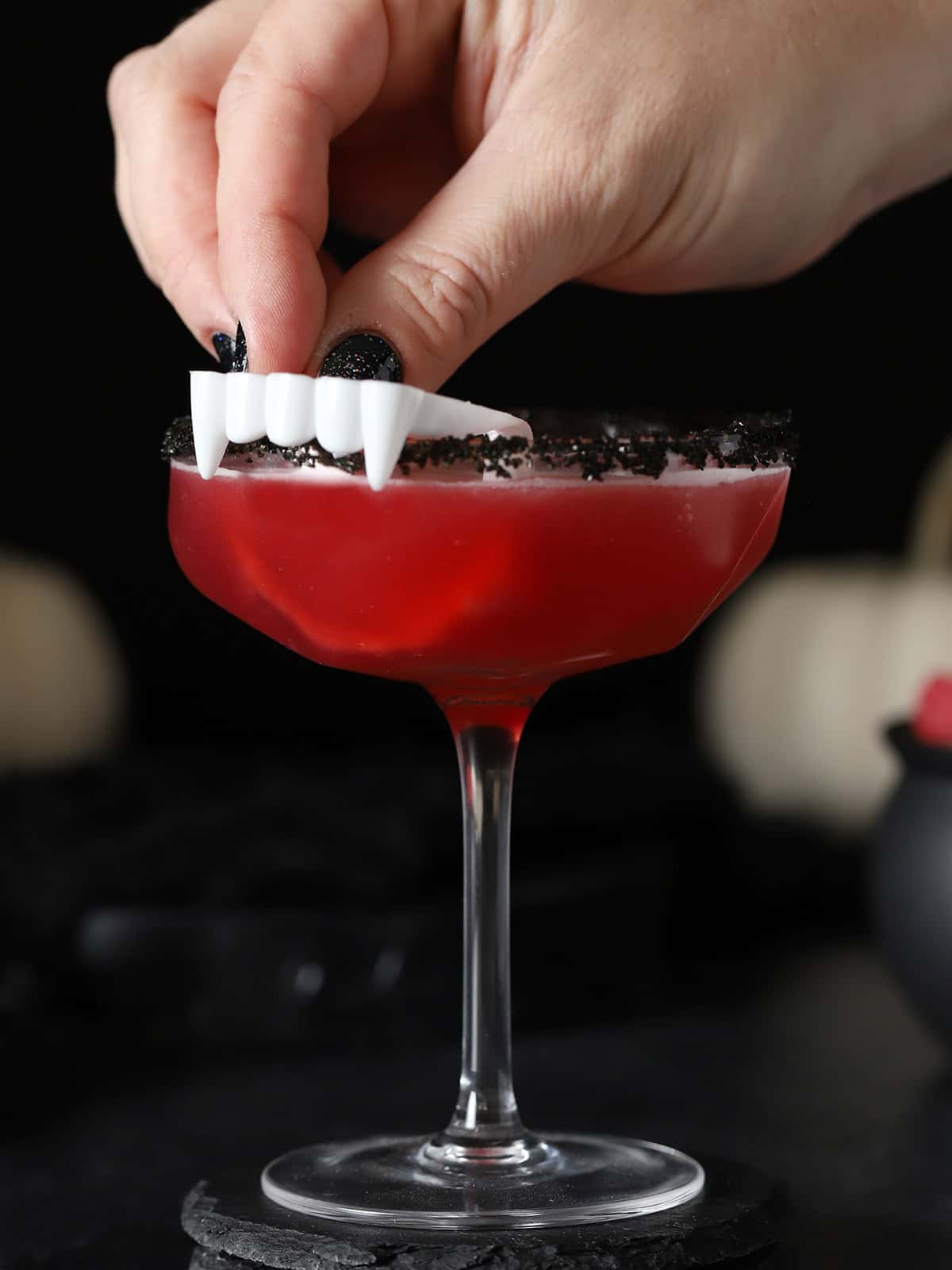 A hand placing a pair of white plastic vampire teeth onto the side of a glass of red Vampire's Kiss cocktail.