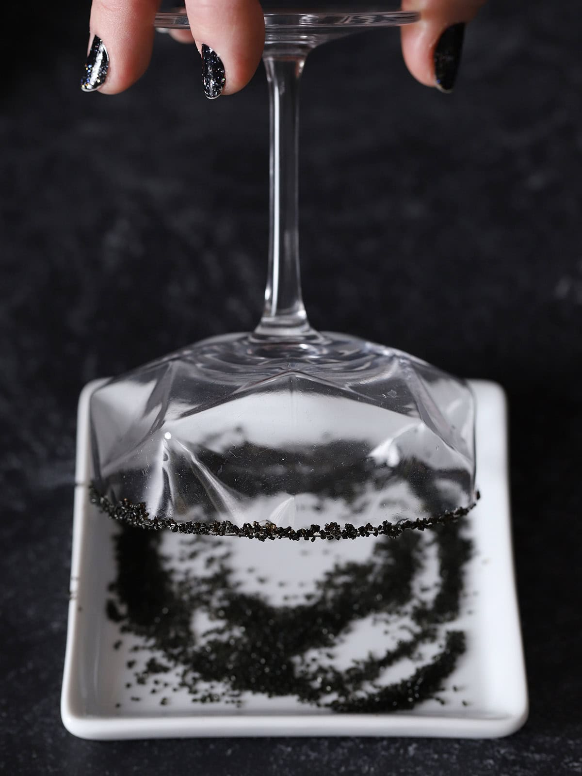 A hand dipping the rim of a coupe glass into a white plate of black sugar.