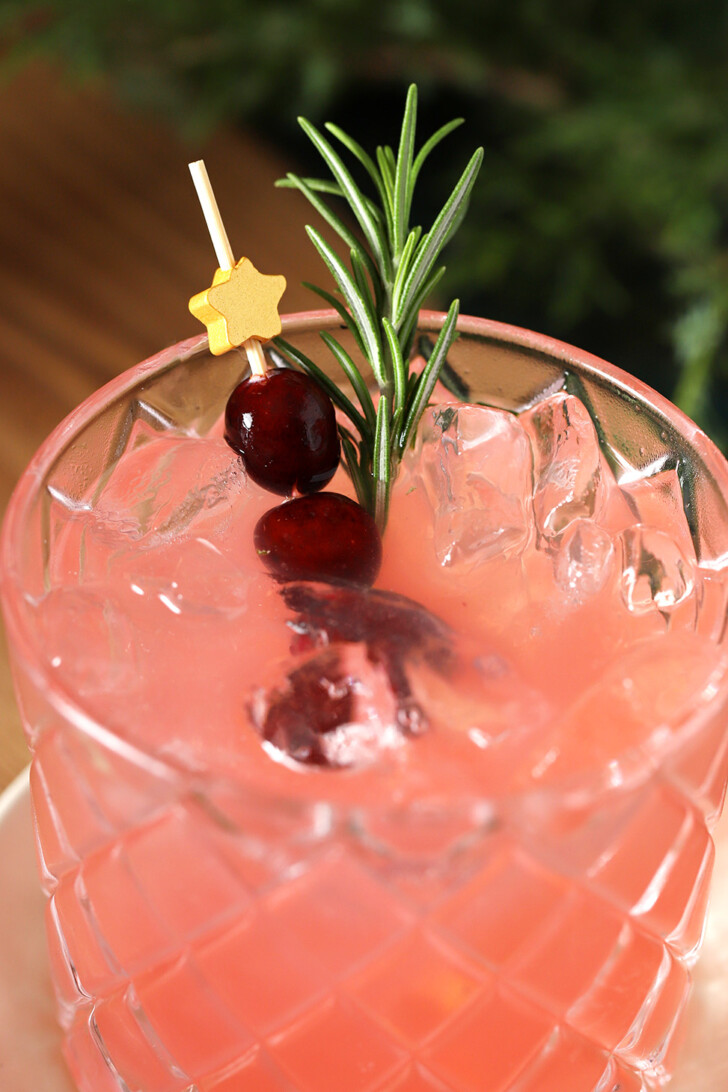 A glass of Cranberry Mocktail garnished with a star cocktail pick with fresh cranberries and a sprig of rosemary.