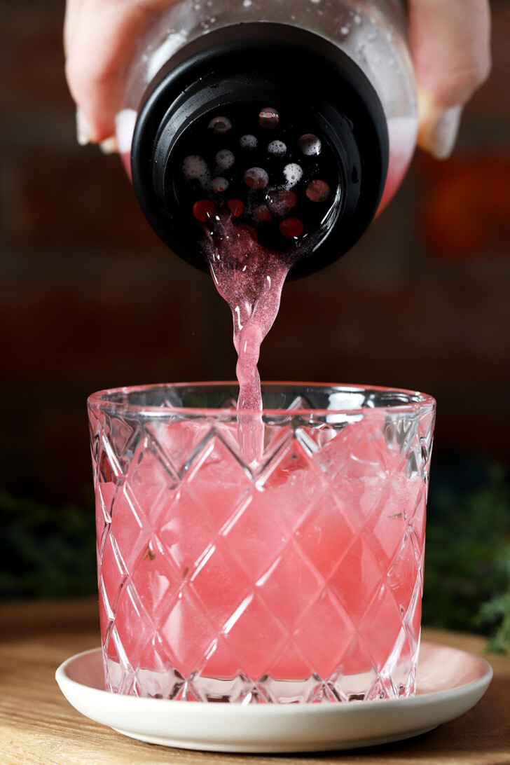 A hand straining cranberry mocktail into a rocks glass filled with ice.