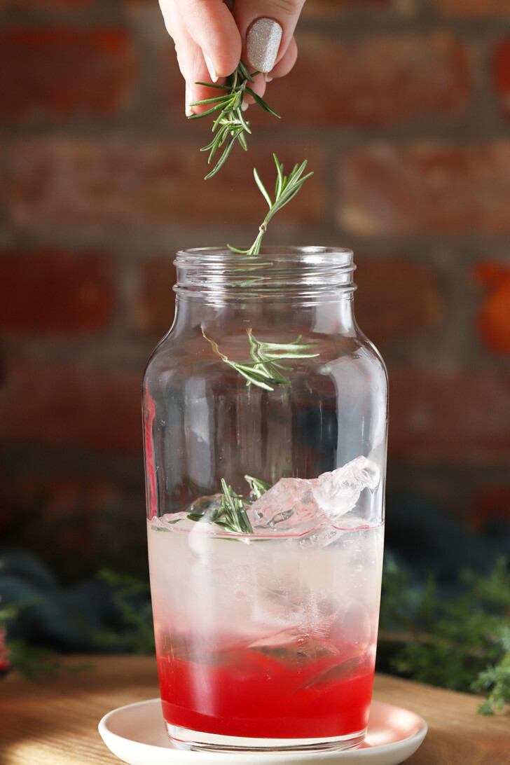 A hand dropping piece of rosemary into a cocktail shaker.