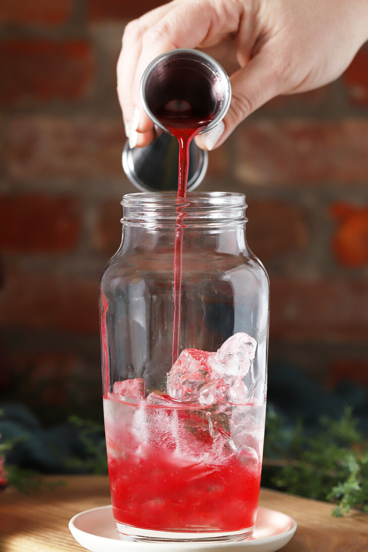 A hand pouring cranberry orange syrup into a cocktail shaker with ice.