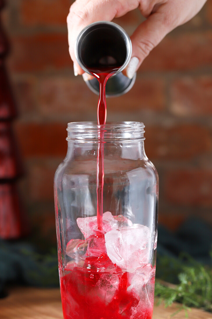 A hand pouring cranberry orange syrup into a cocktail shaker.