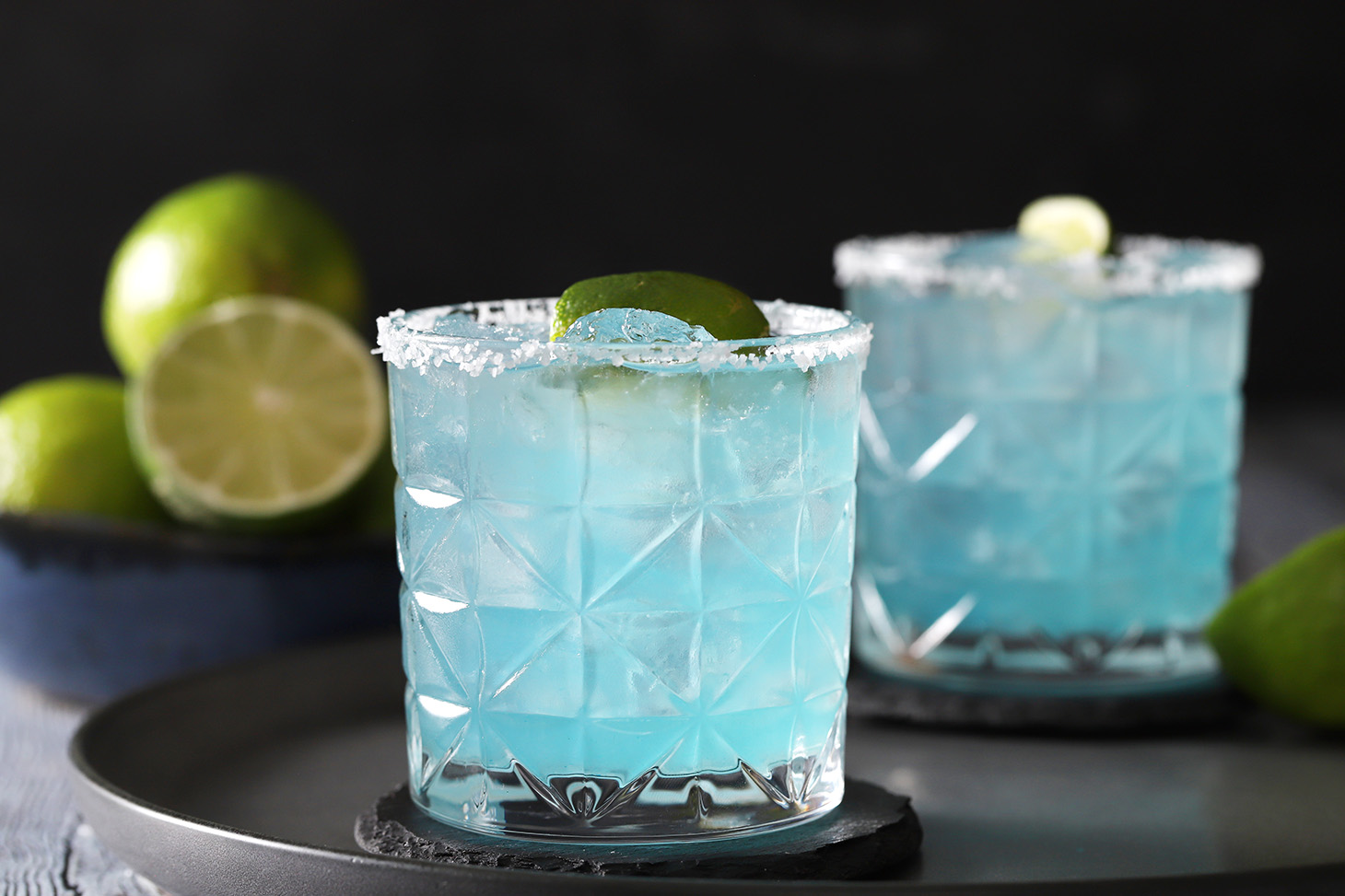 Blue margaritas with salted rims and lime garnishes.