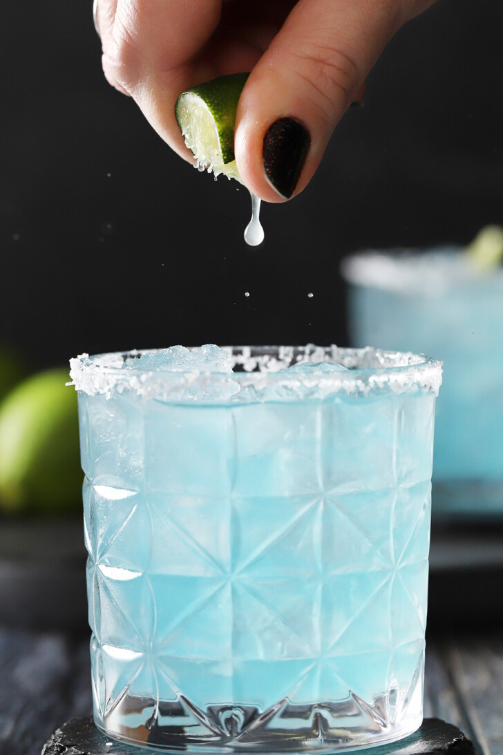A hand squeezing a fresh lime into a a blue margarita.