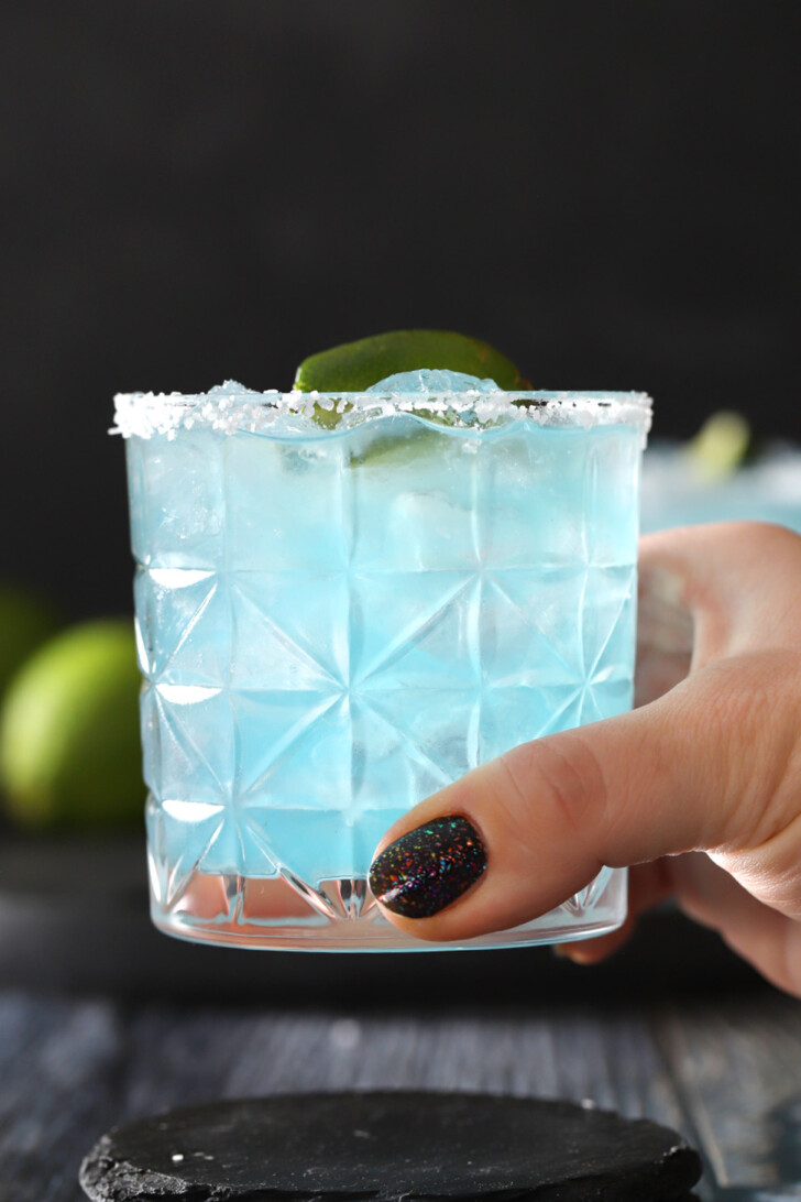 A hand picking up a blue margarita with a salted rim and a lime garnish.