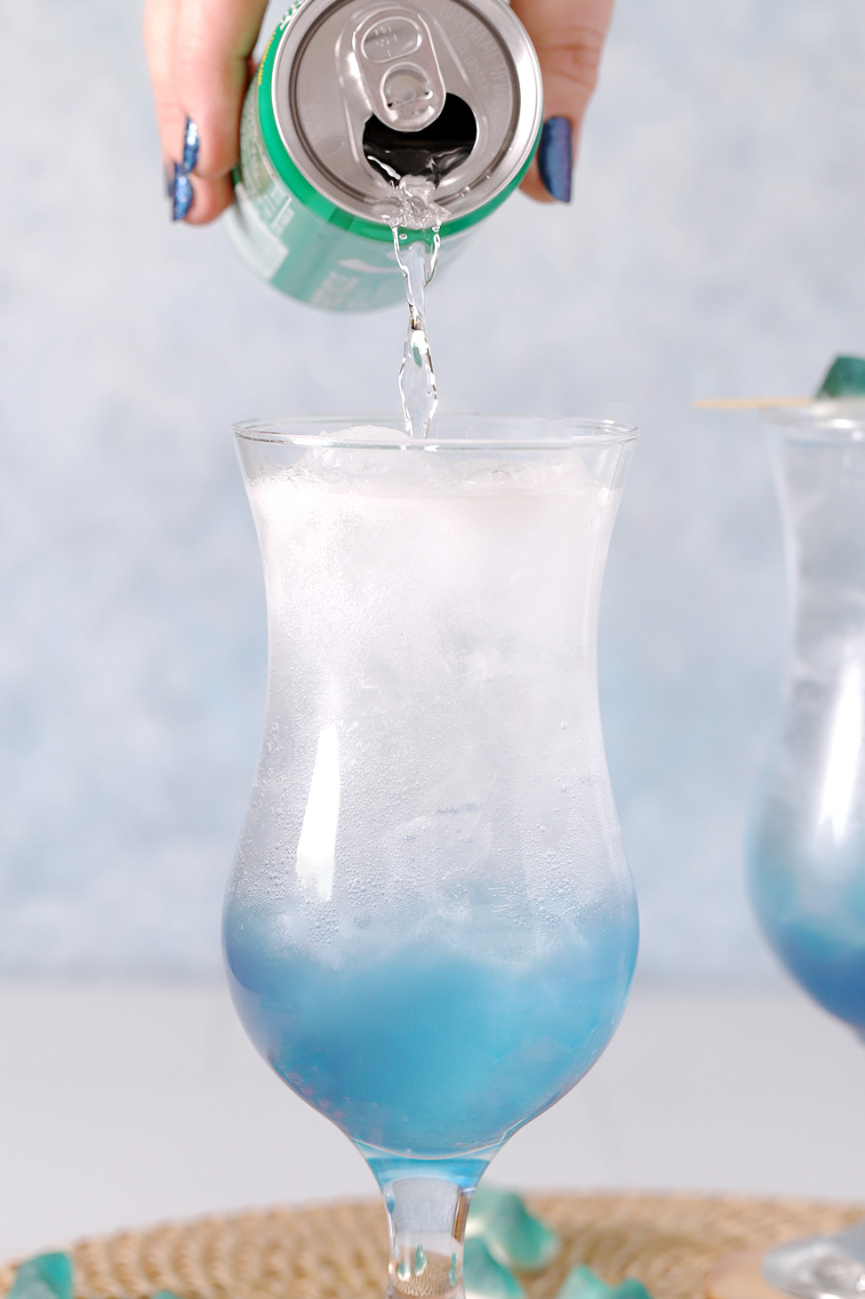 A hand pouring lemon lime soda into a hurricane glass filled with blue mocktail.
