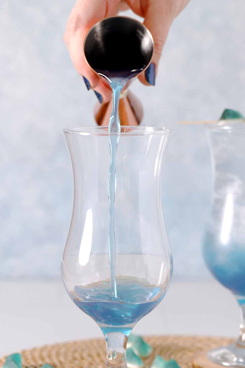 A hand pouring blue curaçao syrup from a jigger into a hurricane glass.