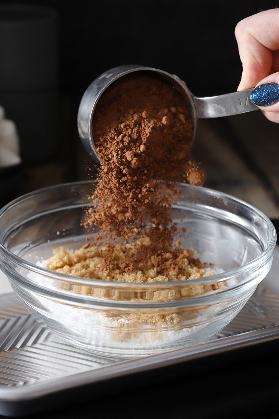 A hand pouring cocoa powder from a measuring cup into a bowl.