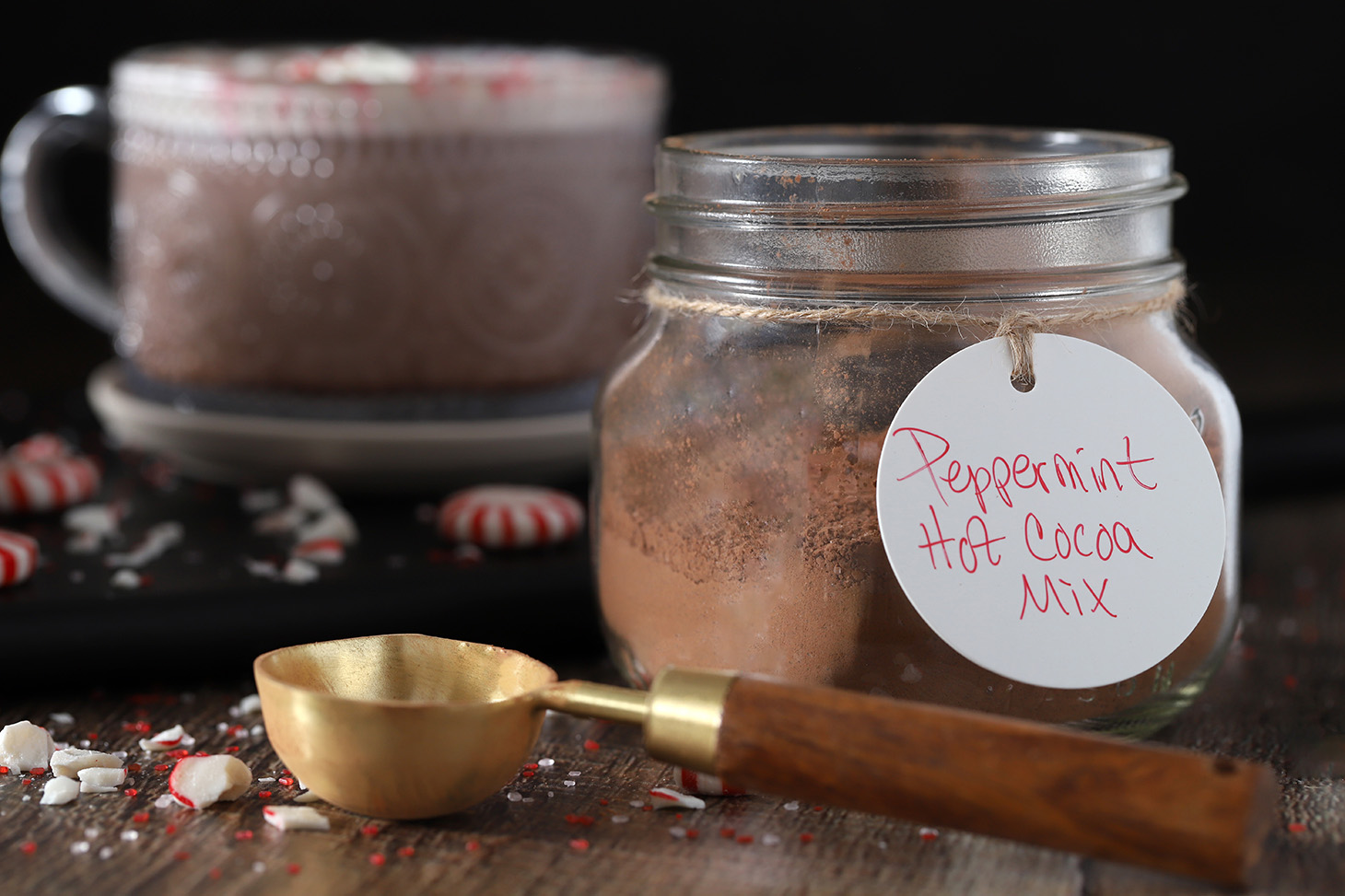 A mason jar filled with peppermint hot cocoa mix with a mug of peppermint hot chocolate in the background.