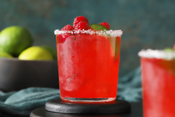 A glass of raspberry margarita sitting on a black coaster, garnished with fresh berries.