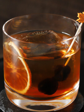 A rocks glass of Maple Old Fashioned cocktail with a dried orange and maple leaf cocktail pick of cherries for garnish.