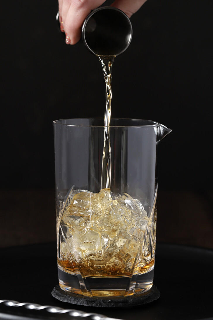 A hand pouring bourbon from a jigger into a mixing glass half filled with ice.