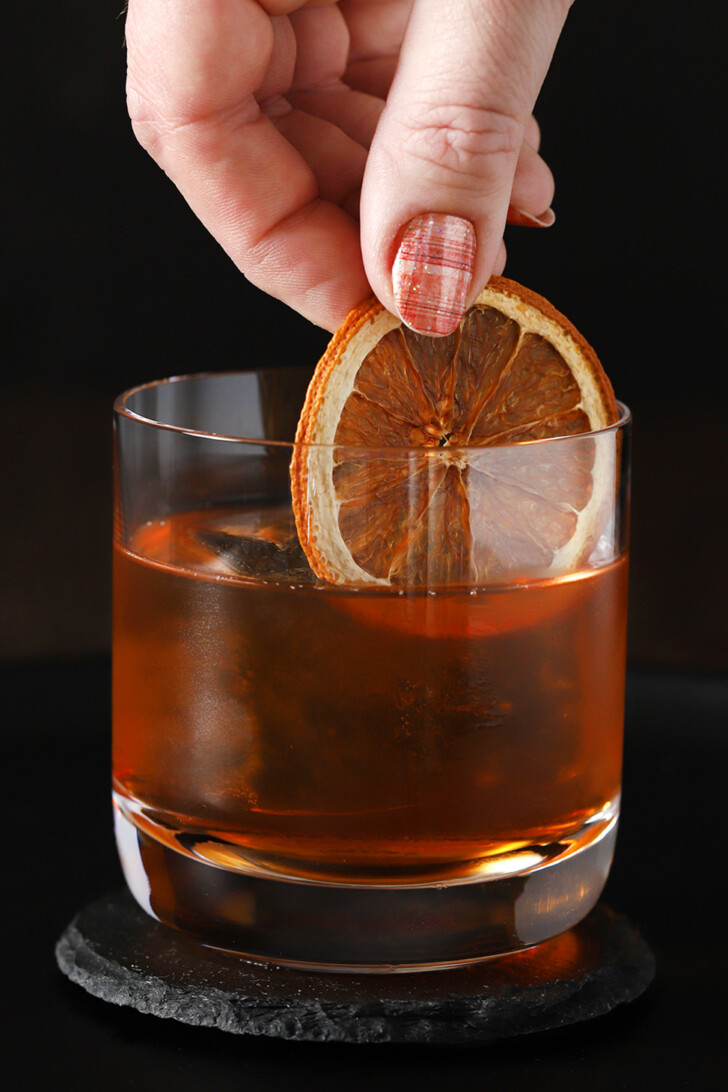 A hand placing a dried orange slice into a rocks glass of maple old fashioned cocktail.