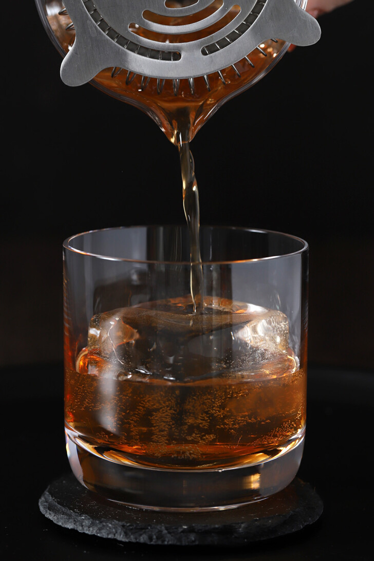 A maple old fashioned cocktail being strained from a mixing glass into a rocks glass and poured over one large, clear ice cube.