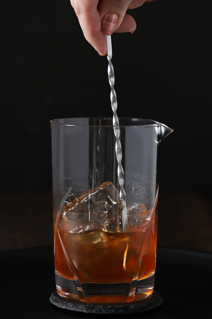 A hand stirring maple old fashioned cocktail in a mixing glass.