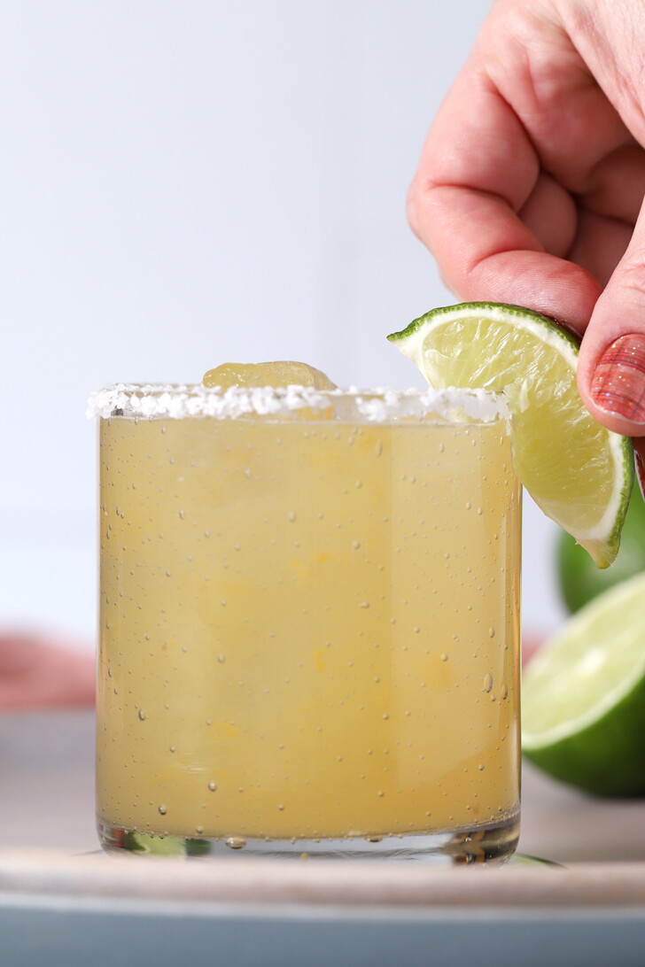 A hand placing a slice of lime onto the edge of the slice of a peach margarita.