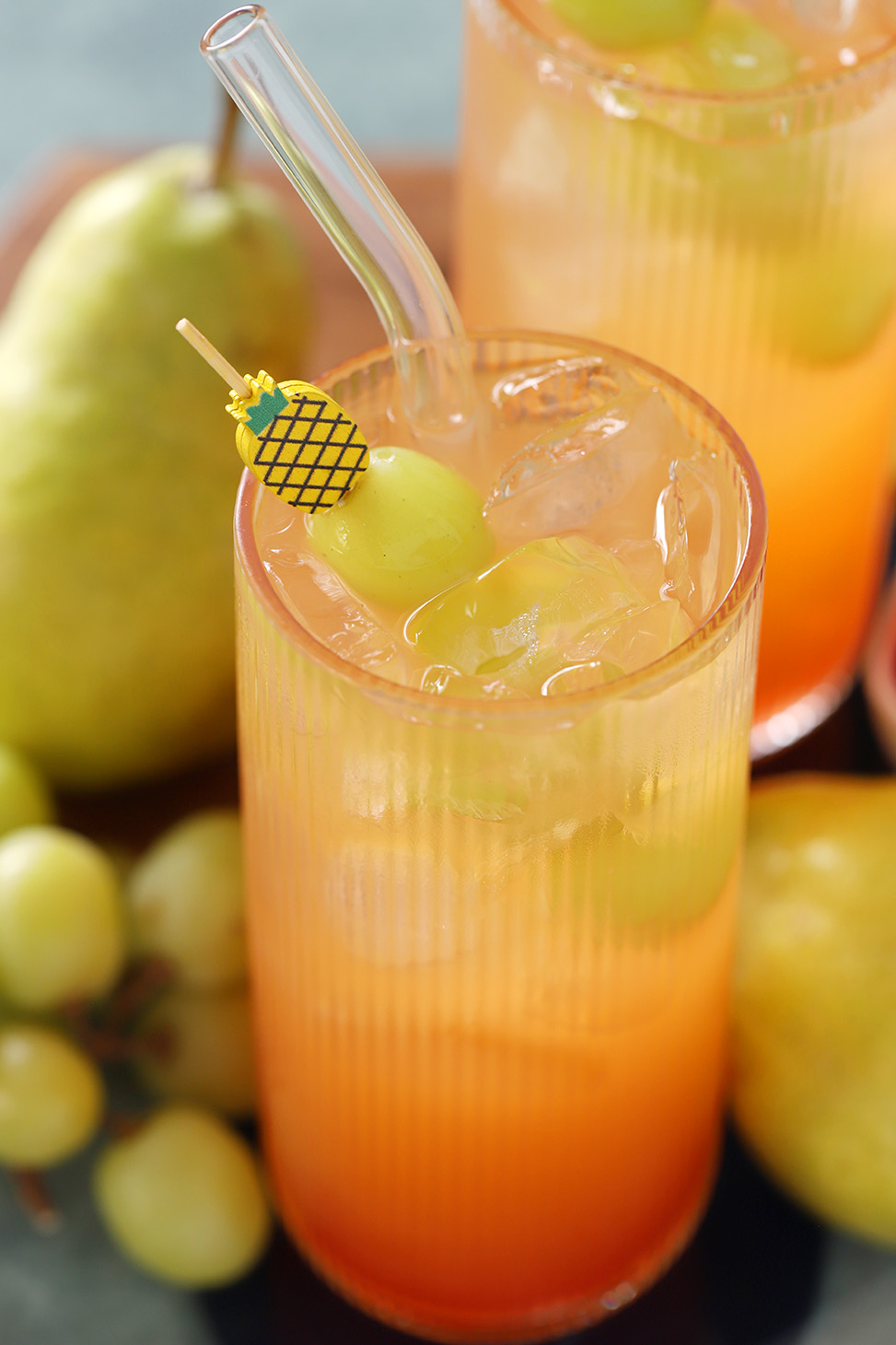 A glass of fruit punch mocktail garnished with green grapes on a pineapple cocktail pick, surrounded by fresh green grapes and pears.