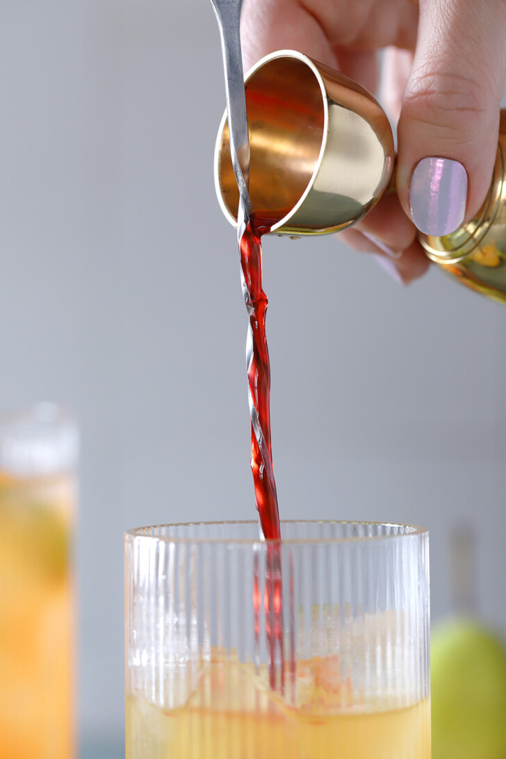 A hand pouring grenadine from a gold jigger down a bar spoon into a glass filled with ice and juice.