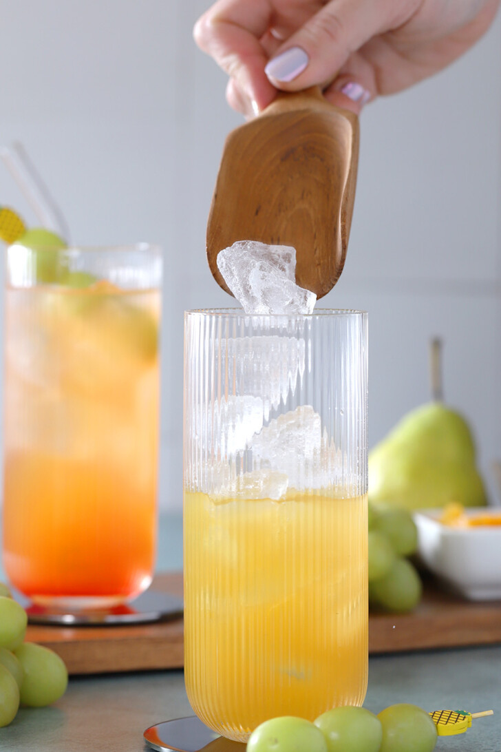 A hand using a wooden scoop to pour ice into a tall glass with juice in it.