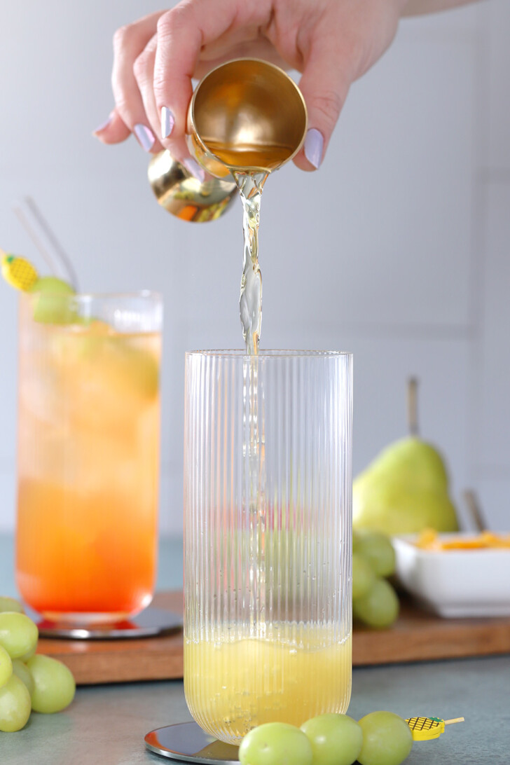 A hand pouring white grape juice from a gold jigger into a tall glass.