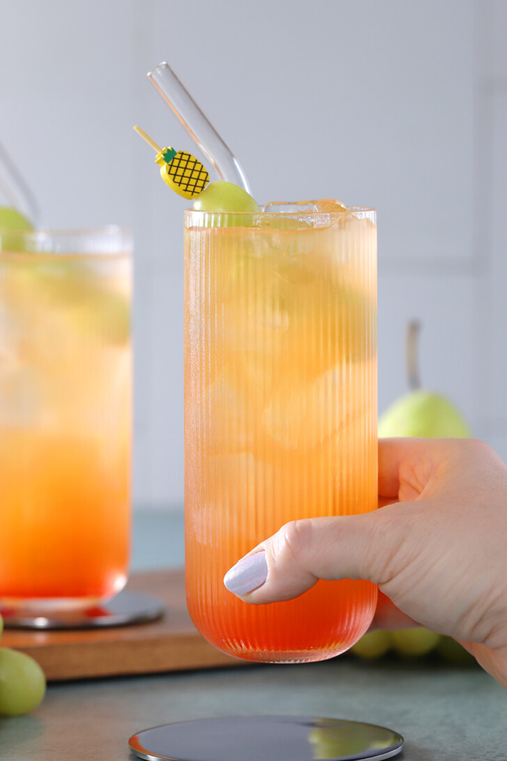 A hand picking up a glass of fruit punch mocktail garnished with green grapes on a pineapple cocktail pick.