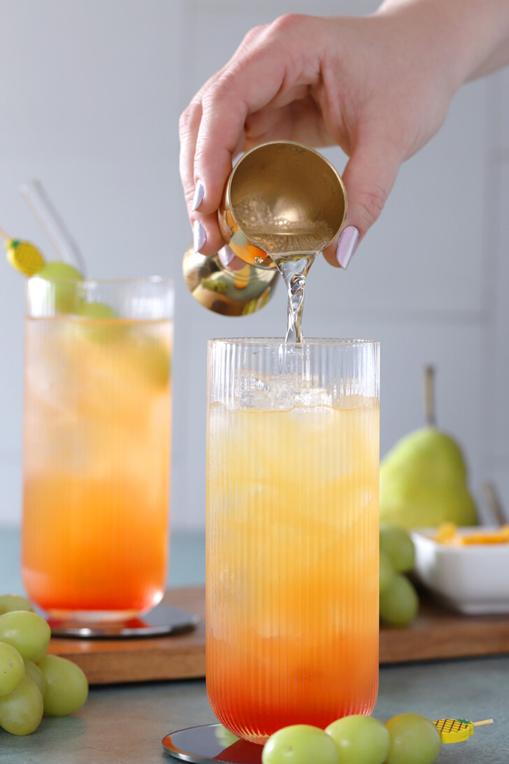 A hand pouring lemon lime soda from a gold jigger into a glass of juice and ice.