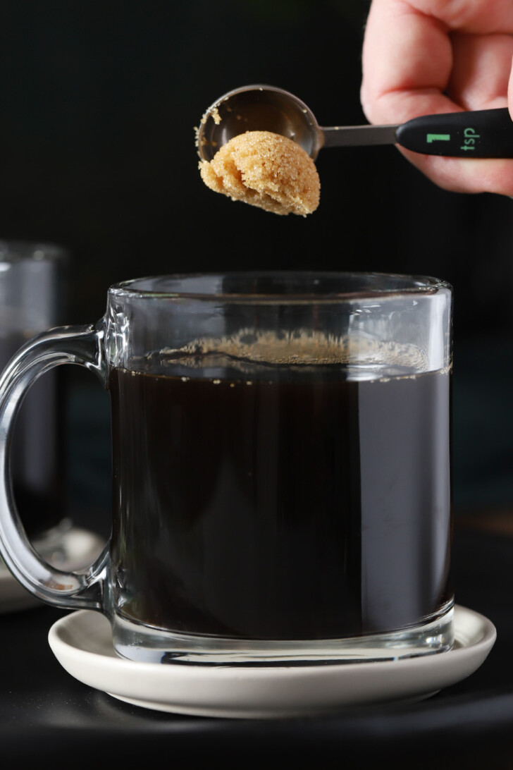 A hand dropping brown sugar from a teaspoon into a glass mug of hot coffee.