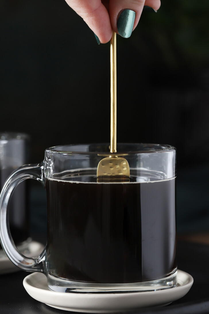 A hand stirring a spoon in a glass cup of coffee.