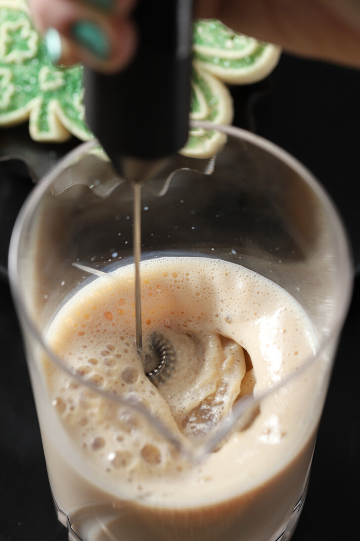 A hand using a milk frother to combine all ingredients together in a mixing glass.