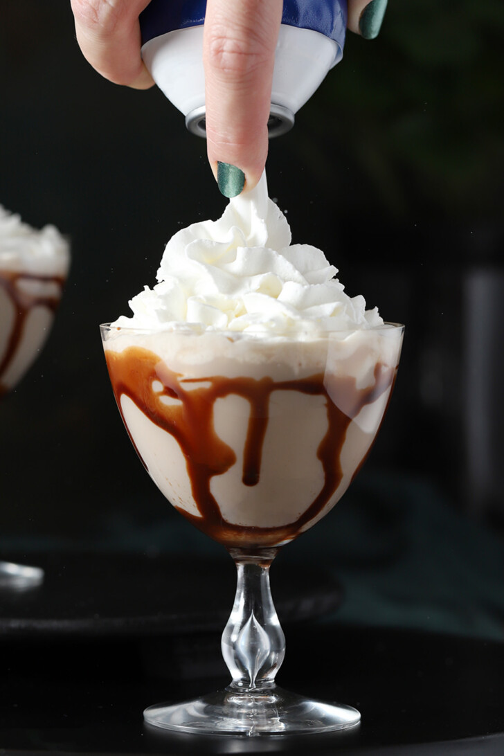 A hand placing a pile of whipped cream on top of a glass of Irish cream cocktail in glasses swirled with chocolate sauce.