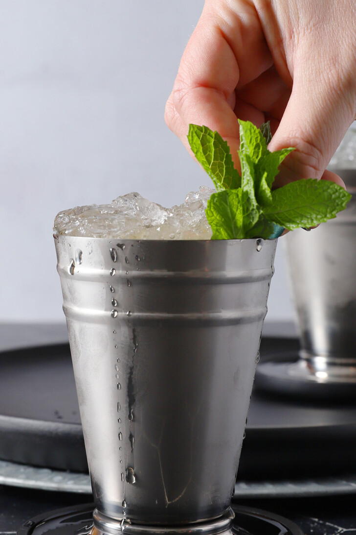 A hand placing a large sprig of mint into a silver cup of mint julep cocktail.