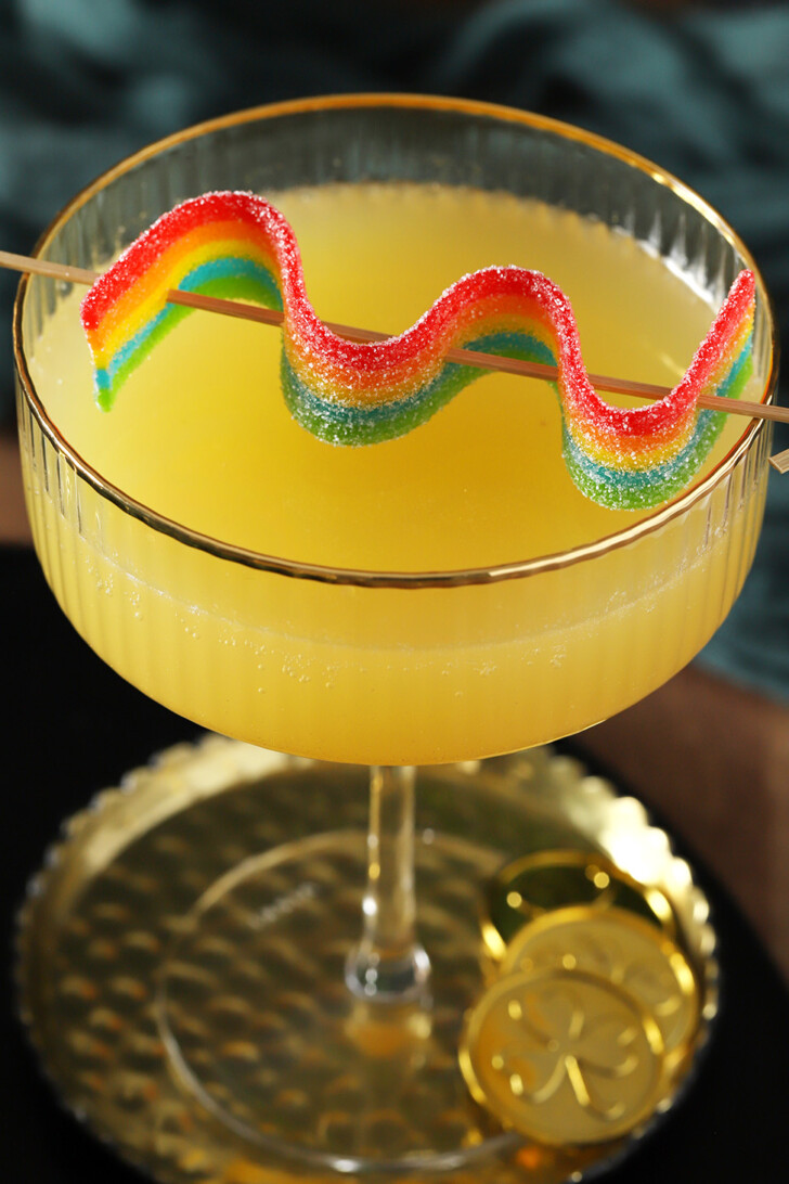 A yellow Pot of Gold St. Patrick's Day mocktail in a coupe glass with a wavy rainbow candy on a cocktail pick set on top of the glass.