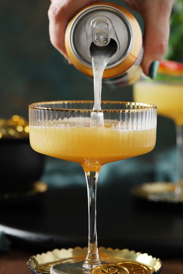 A hand pouring ginger beer into a coupe glass filled with pot of gold mocktail.