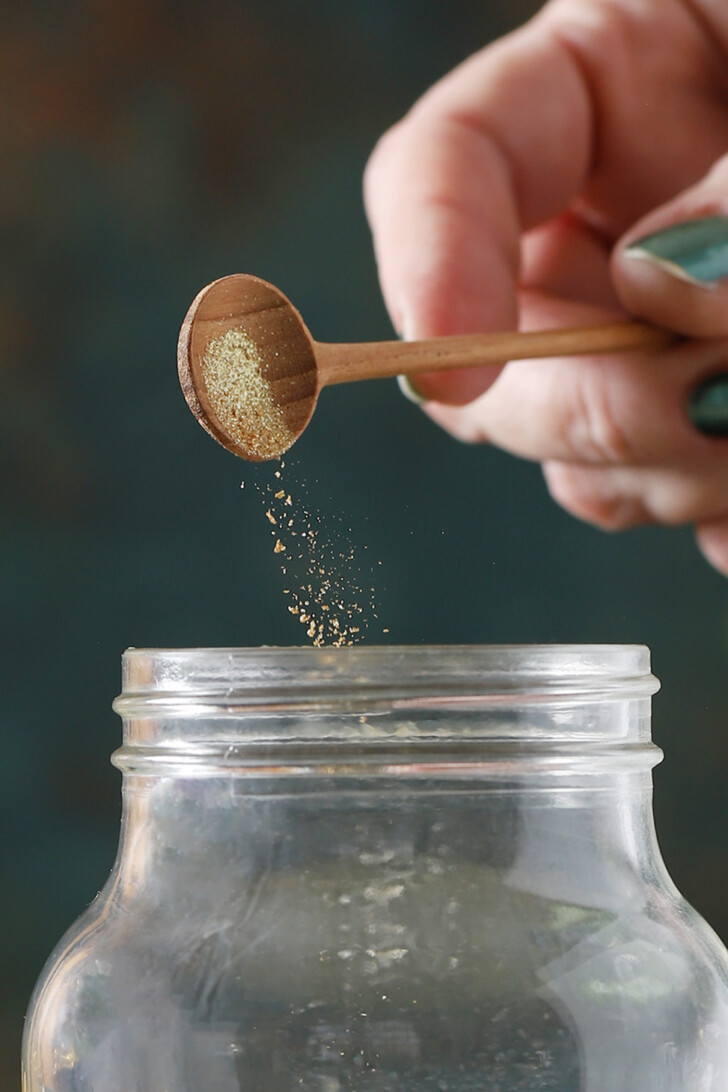 A hand dropping edible glitter from a wooden spoon into a cocktail shaker.