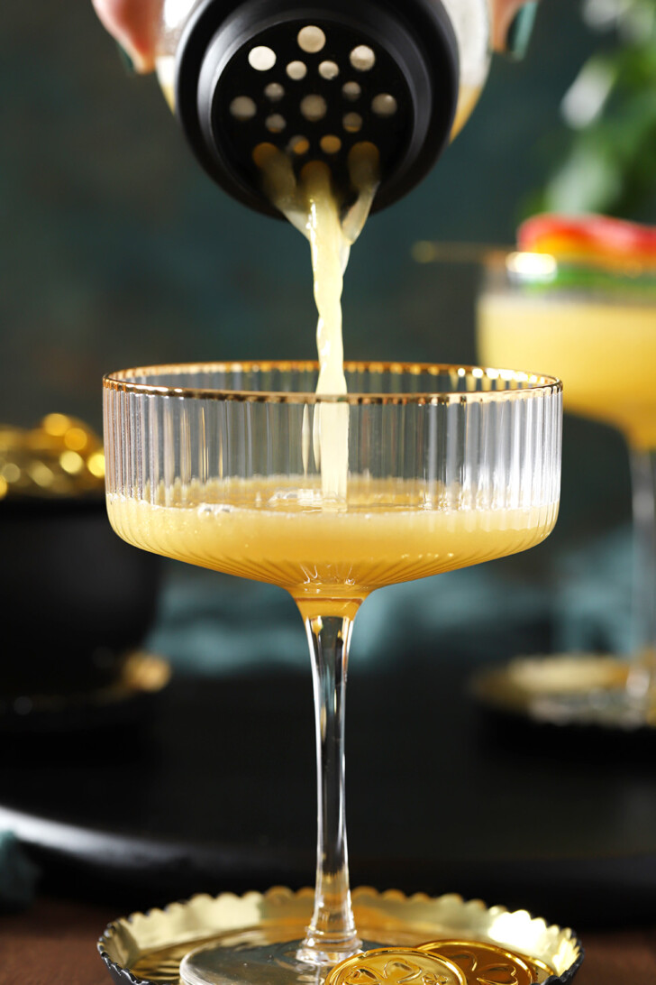 A yellow Pot of Gold mocktail being poured from a cocktail shaker into a coupe glass.