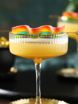 A yellow Pot of Gold St. Patrick's Day mocktail in a coupe glass with a wavy rainbow candy on a cocktail pick set on top of the glass.
