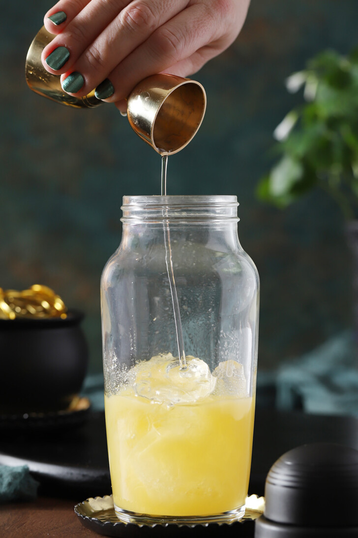 A hand pouring simple syrup from a jigger into a cocktail shaker.