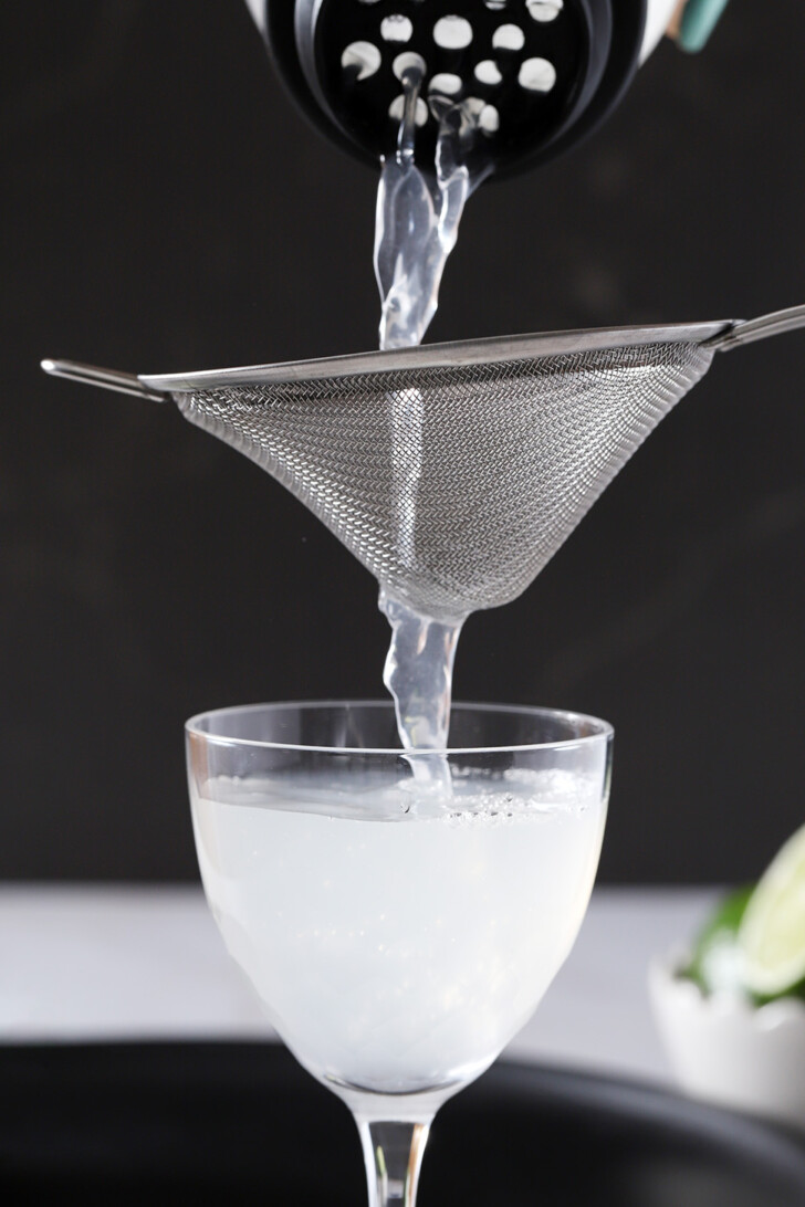 Gin gimlet being poured from a cocktail shaker, through a fine mesh strainer and into a Nick and Nora glass.