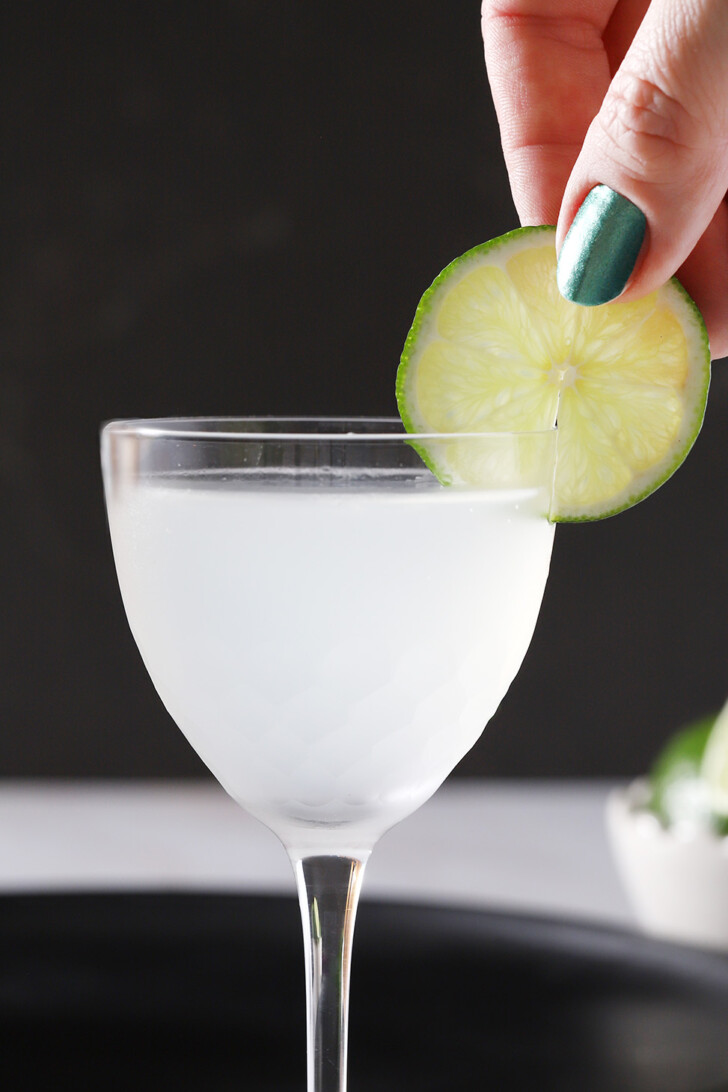 A hand placing a lime wheel onto the side of a Nick and Nora glass filled with gin gimlet.